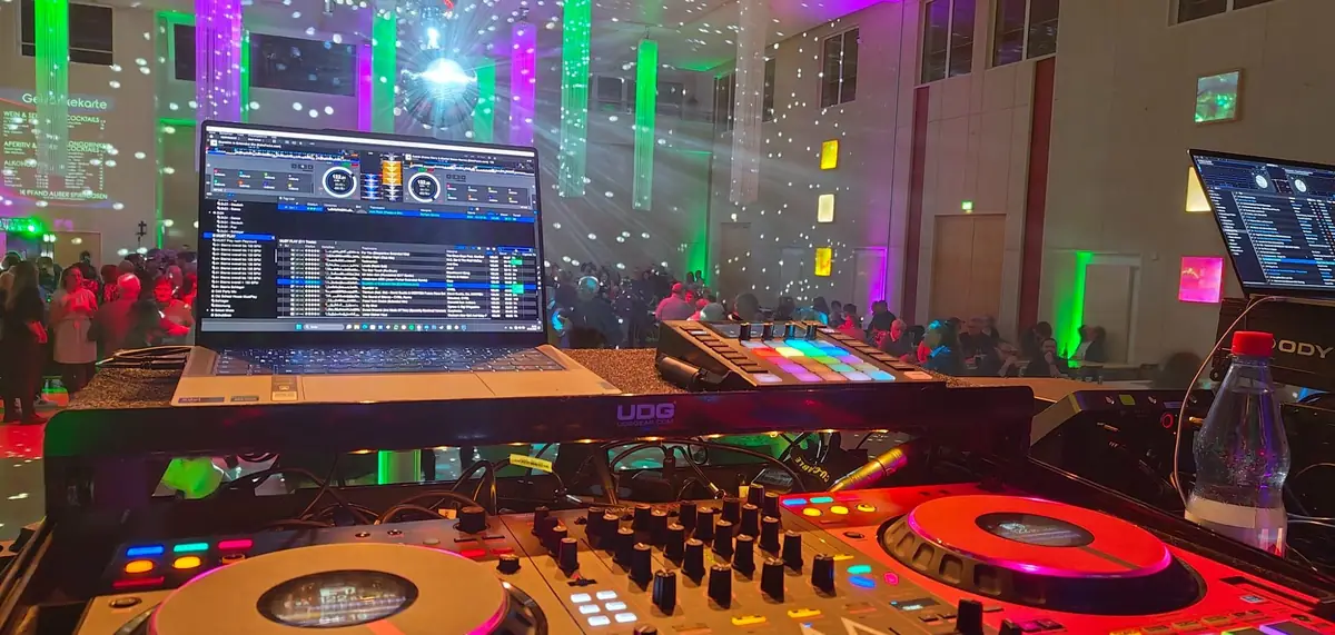 DJ-Setup mit Mischpult, bunten Lichtern und einer tanzenden Menge im Hintergrund.