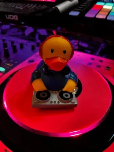Ein gelber Gummiente als DJ an einem Plattenteller mit bunten Lichtern im Hintergrund.