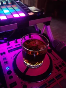 Ein Glas mit Getränk steht auf einem DJ-Pult, umgeben von bunten Lichtern. Photo by DJ Mirko Anders